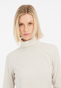 Pull à col roulé beige clair, côtelé, avec des manches longues, offrant une coupe ajustée et un tissu texturé pour la chaleur et le confort.