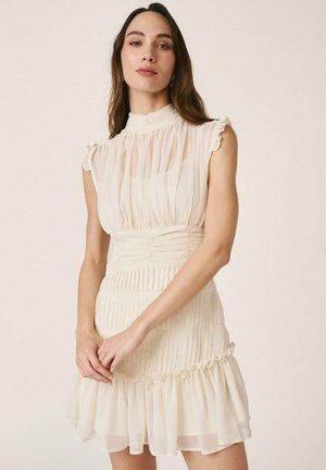 Mujer con vestido crema plisado sin mangas, con cuello alto y bajo con volantes, de pie con las manos entrelazadas sobre un fondo claro.