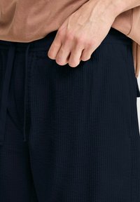 Navy blue textured shorts with a ribbed fabric pattern, featuring an adjustable drawstring waist. A hand grips the waistband.