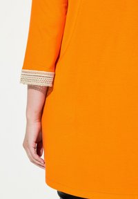 Leuchtend orangene Tunika mit Dreiviertelärmeln, die einen dekorativen Spitzeabschluss an den Ärmelenden aufweist. Weicher Stoff und gerader Saum.