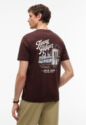 Mann trägt ein dunkelbraunes Tommy Hilfiger T-Shirt mit New-York-Brücke- und Skyline-Grafik, beige Hose und eine Armbanduhr, von hinten gesehen.