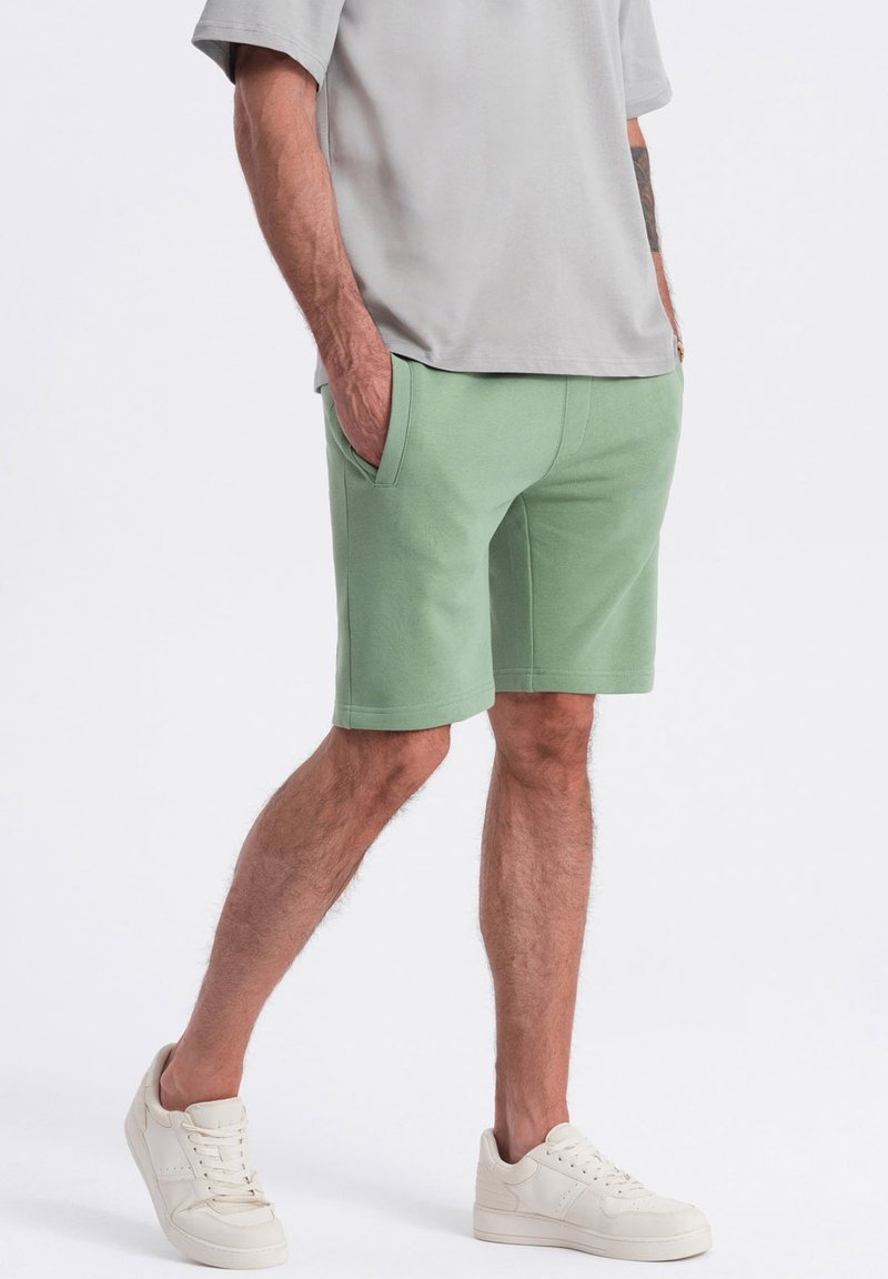 Shorts vert clair en tissu doux, dotés de poches latérales et d'une coupe décontractée. Portés avec un t-shirt gris et des baskets blanches.