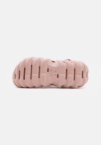 Pink molded foam footwear with a wavy texture and circular cutouts, featuring a flat sole design and marked branding on the side.