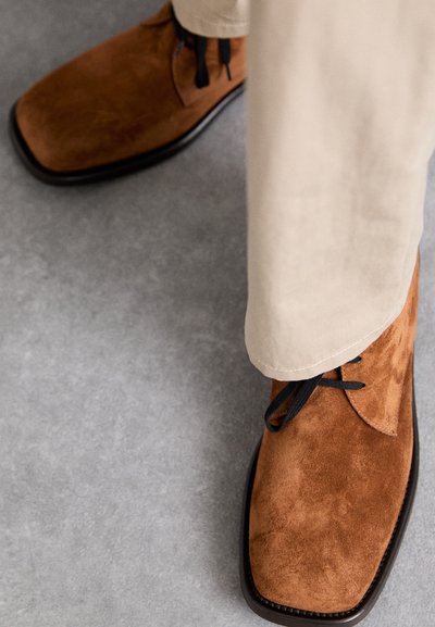 Zapatos de ante marrón con punta cuadrada y cordones negros; acompañados de pantalones beige claros, reposando sobre una superficie gris.