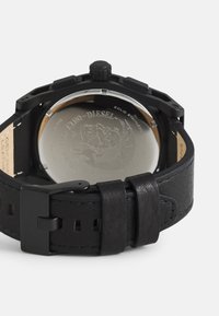 Schwarzlederarmbanduhr mit einem soliden Edelstahlboden, ausgestattet mit einem strukturierten Armband, einer Metallschnalle und einem ausgeprägten seitlichen Krone-Detaill.