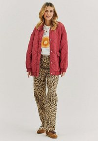 Červená prešívaná bunda cez grafické tričko, doplnená béžovými nohavicami s leopardím vzorom a hnedými topánkami. Oblečenie má voľný strih a neformálny dizajn.