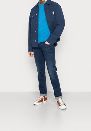 Hombre con chaqueta azul oscura, camiseta azul, jeans oscuros y zapatillas altas marrones de pie contra un fondo blanco.