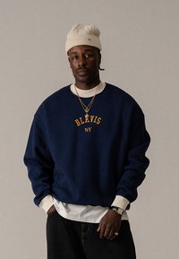 Tengerészkék pulóver sárga "BLKVIS NY" felirattal, fehér ing fölött viselve, fekete farmerrel és krémszínű kötött sapkával párosítva.