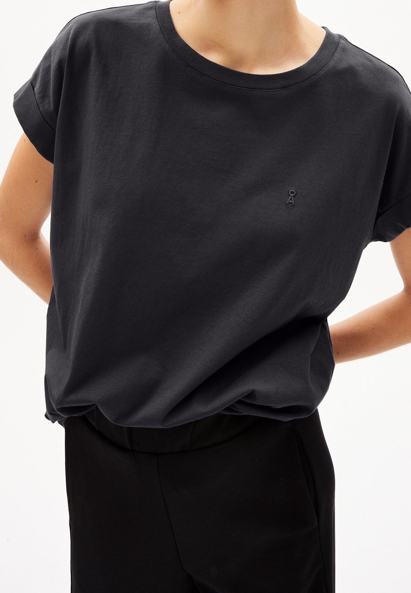Personne portant une chemise noire ample à manches courtes avec un petit logo brodé sur la poitrine et un pantalon noir, les mains dans le dos.