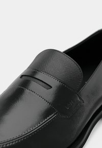 Mocassin en cuir noir à finition lisse, avec une bride texturée et un détail de coutures discrètes. Logo embossé sur le côté.