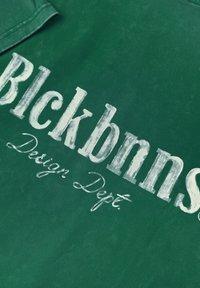 Donkergroen stoffen t-shirt met witte tekst: "Blckbnns" in vette letters en "Design Dept." in een cursieve stijl. Zacht, gestructureerd materiaal.
