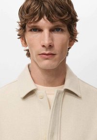 Giacca camicia beige chiaro con colletto, realizzata in tessuto morbido, caratterizzata da una chiusura con bottoni e silhouette dritta, indossata sopra un maglione chiaro.