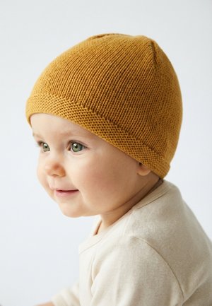 Lächendes Baby mit grünen Augen, das eine senfgelbe Strickmütze und ein cremefarbenes Shirt trägt, nach links blickend vor weißem Hintergrund.