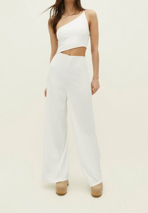 Vrouw die een witte crop top met één schouder en uitsparing draagt, en witte, hooggetailleerde, wijde broekspijpen tegen een effen lichte achtergrond.