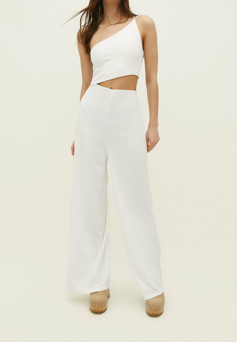 Femme portant un crop top blanc asymétrique avec découpes et un pantalon blanc taille haute à jambes larges sur un fond clair uni.