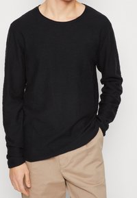 Svart långärmad tröja med rund halsringning och ett mjukt, texturerat tyg. Bärs med ljusare byxor i en avslappnad miljö.