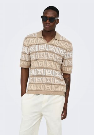 Homme portant des lunettes de soleil noires, une chemise en maille beige à motifs et manches courtes et un pantalon clair texturé, debout avec une main dans la poche.