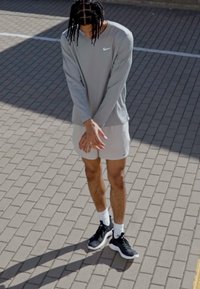 Svetlo šedá dlhá rukávová košeľa Nike, biele kraťasy, čierne športové topánky s bielymi podrážkami, stojace na vzorovanej dlažbe.