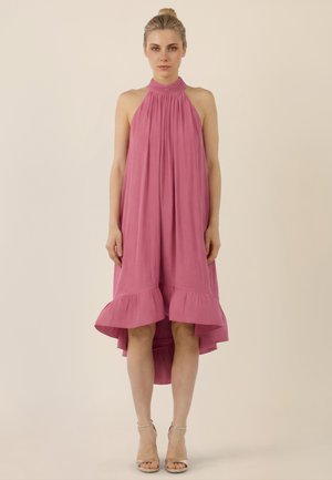 Kobieta stojąca wyprostowana, ubrana w różową, bezrękawnikową sukienkę z wysokim kołnierzem, luźno opadającą, z falbankowanym dołem oraz cielistymi szpilkami na kostkę, na beżowym tle.
