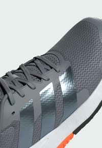 Zapatilla deportiva gris con parte superior de malla, acentos reflectantes, detalles texturizados y suela blanca con un acento naranja.