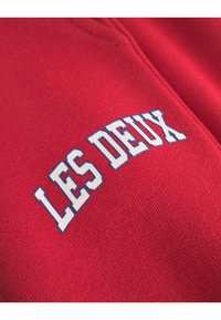 Crvena tkanina s glatkom teksturom sadrži natpis "LES DEUX" u podebljanim bijelim slovima s plavim naglaskom. Slova imaju geometrijski stil.