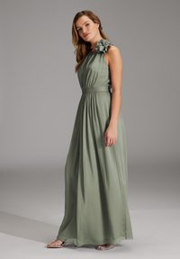 Vrouw staande in een lang, zandgroen, mouwloos jurkje met een gerimpelde schouderdetails en zilveren sandaaltjes tegen een grijze achtergrond.