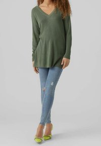 Modelka v dlouhém, zeleném svetrém s výstřihem do V pózuje v potrhaných světlých džínách a jasně žlutých podpatcích na neutrálním pozadí.