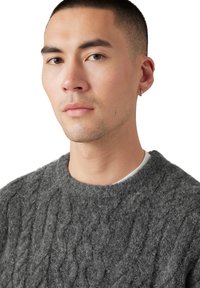 Suéter gris de punto con un patrón de trenzas, escote redondo y apariencia suave y texturizada. La modelo tiene el cabello corto y lleva un aro pequeño en una oreja.