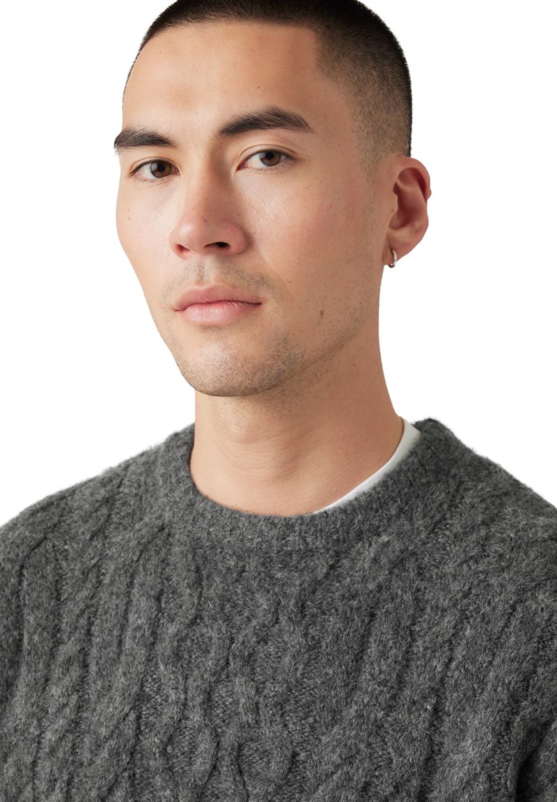 Suéter gris de punto con un patrón de trenzas, escote redondo y apariencia suave y texturizada. La modelo tiene el cabello corto y lleva un aro pequeño en una oreja.