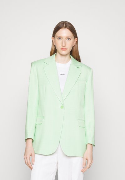 Blazer vert menthe avec deux poches avant, fermeture à un seul bouton et texture visible. Porté par-dessus un t-shirt blanc et associé à un pantalon blanc.