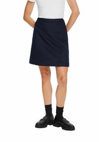 Femme portant une jupe bleu marine mi-longue, un haut blanc, des chaussettes noires et des mocassins noirs, debout les mains derrière le dos.