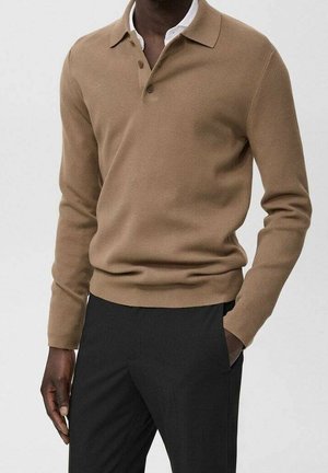 Mann trägt ein beiges Langarm-Poloshirt über einem weißen Hemd mit Kragen, kombiniert mit schwarzen Hosen, Hand in der Tasche.