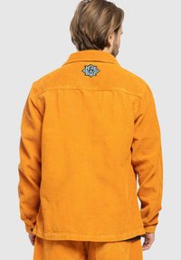 Orange corduroy jacket with a back patch in black featuring geometric design. Regular fit with buttoned cuffs and straight hem.