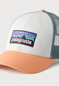 Καπέλο τύπου trucker σε λευκό και πορτοκαλί χρώμα με πλέγμα στο πίσω μέρος και πολύχρωμο έμβλημα λογότυπου Patagonia στην μπροστινή πλευρά.