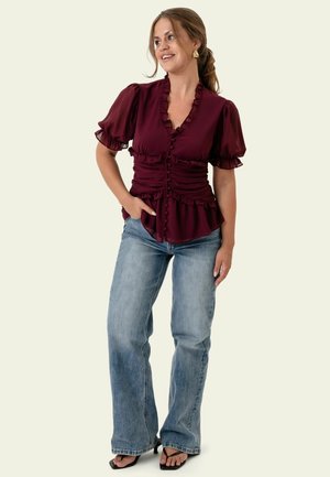 Femme portant un chemisier bordeaux à volants avec manches bouffantes, jean bleu clair, sandales noires à talons et boucles d'oreilles en or, debout avec une main dans la poche.