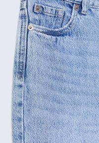 Primer plano de unos vaqueros de mezclilla azul claro que muestra el bolsillo delantero, el bolsillo para monedas, el lazo para el cinturón y los remaches de cobre sobre una tela texturizada.
