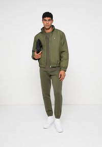 Ελαιώδης πράσινος μπόμινγκ τζάκετ με φερμουάρ μπροστά, ριμπέ κοψίματα στους καρπούς και τη μέση. Φορεμένο πάνω από ένα αντίστοιχο ελαιώδες t-shirt και φόρμες, με λευκά αθλητικά παπούτσια.