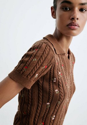 Vrouw draagt een bruin gebreid T-shirt met korte mouwen en kleine geborduurde bloemen, vanaf schouder tot kin afgebeeld tegen een effen achtergrond.