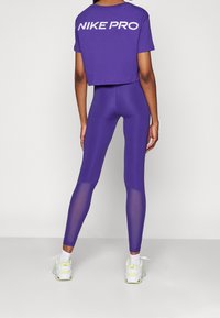 Fioletowy krótki t-shirt z nadrukiem "NIKE PRO" w kolorze białym, zestawiony z fioletowymi legginsami z siateczkowymi detalami na dolnych nogawkach oraz białymi trampkami.