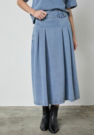 Woman wearing a textured blue midi denim skirt, matching top, and black heeled ankle boots, standing on a concrete floor.