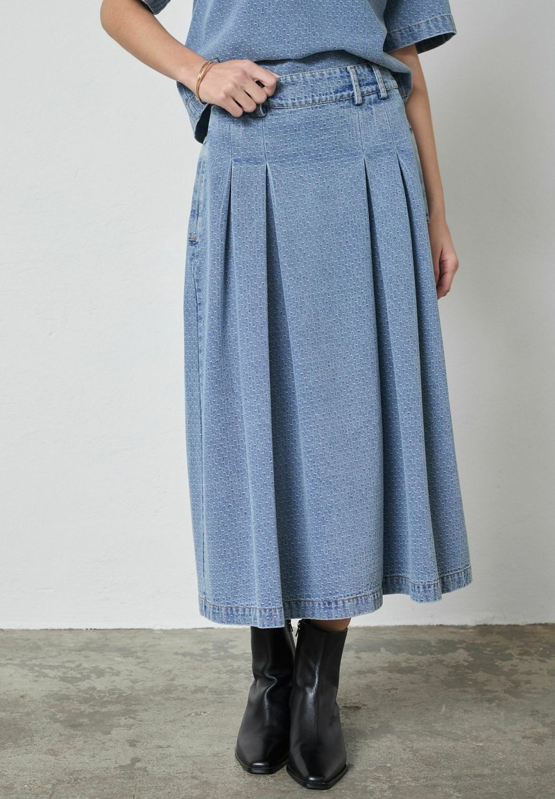 Nő texturált kék midi farmer szoknyában, hozzáillő felsőben és fekete magassarkú bokacsizmában, betonpadlón állva.