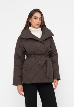Femme portant une veste matelassée sombre avec ceinture sur un pull à col roulé blanc et un pantalon noir, debout devant un fond clair uni.