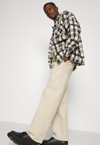 Camisa de flanela xadrez em preto e branco por cima de um suéter verde, combinada com calças largas em tom creme e sapatos pretos, de pé contra um fundo neutro.