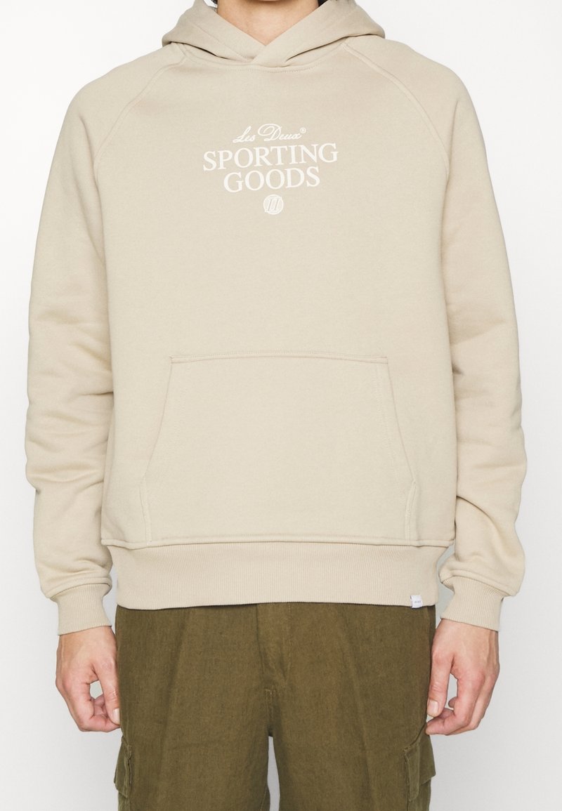 Beige hoodie gemaakt van zachte stof, met een kangoeroezak, geribbelde manchetten en zoom, en het logo "SPORTING GOODS" in witte letters op de voorkant.