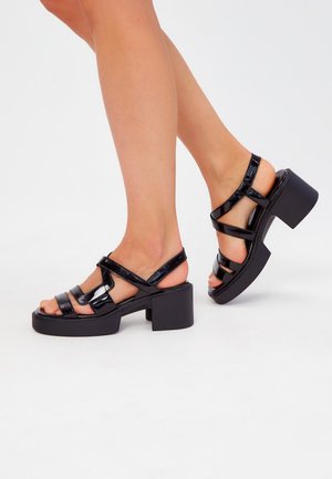 Sandali in vernice nera con tacco quadrato, caratterizzati da più cinturini incrociati e un design con punta aperta. Tacco spesso per una maggiore stabilità.