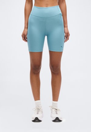 Spodnji del telesa osebe, ki nosi svetlo modre športne kratke hlače z visokim pasom, bele nogavice in bele tekaške copate na enobarvnem ozadju.