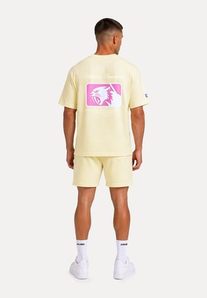 Mann steht mit dem Rücken zur Kamera, trägt hellgelbe Shorts und ein T-Shirt mit einer pinkfarbenen, brüllenden Tiger-Grafik auf dem Rücken. Weiße Socken und Turnschuhe.
