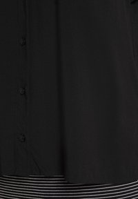 Camicia nera con bottoni, orlo sopra un tessuto a righe orizzontali bianche e nere, che mostra il dettaglio inferiore anteriore e i bottoni.