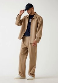 Tijgerkleurig getailleerd jack, navy T-shirt en beige broek met wijde pijpen. Witte sneakers. Man past een navy baseballpet met logo aan. Gladde stof, ontspannen pasvorm.