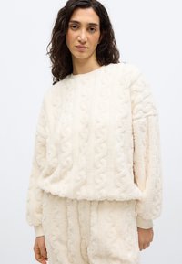 Γυναίκα που φοράει χνουδωτό πουλόβερ σε κρεμ απόχρωση με υφή και ταιριαστό παντελόνι, στέκεται μπροστά σε απλό λευκό φόντο.